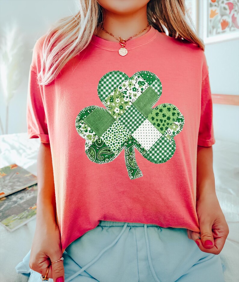 May include: A coral-colored t-shirt featuring a large, patchwork shamrock design. The shamrock is composed of various green patterned fabrics, including gingham, floral, and paisley prints. The shirt has short sleeves and a crew neck, perfect for St. Patrick's Day.