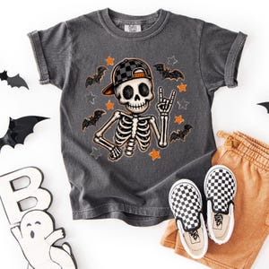 May include: A charcoal gray short-sleeve t-shirt featuring a graphic of a skeleton wearing a black and gray checkered baseball cap. The skeleton is making a hand gesture surrounded by orange and gray stars and black bat silhouettes.