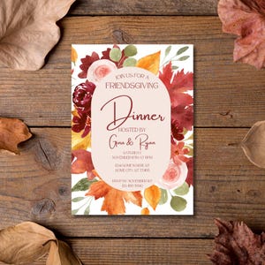 Op de afbeelding: Een Friendsgiving-dineruitnodiging met een bloemendesign. De uitnodiging heeft een crèmekleurige ovaal met de woorden "Dinner" en "Hosted by Gina & Ryan" in een scriptlettertype. De uitnodiging is omgeven door herfstbladeren en bloemen.
