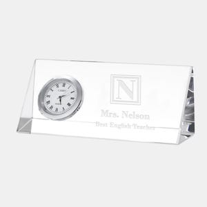 May include: A clear, rectangular desk clock with a silver-colored clock face and Roman numerals. The clock has the text "CAMRY" and "QUARTZ" on the face. The clock also has the engraved text "Mrs. Nelson Best English Teacher" and a letter "N".