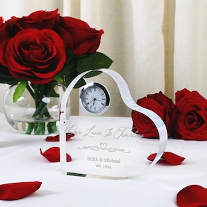 May include: A clear glass heart-shaped clock with the inscription "Our Love Is Timeless" and the names "Edith & Michael" and the date "est. 1994". The clock is surrounded by red rose petals.