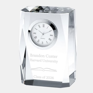 May include: A clear crystal desk clock with a quartz movement. The clock face has Roman numerals and a silver bezel. The crystal is engraved with the name "Brandon Custer", "Harvard University", a book graphic, and "Class of 2026".