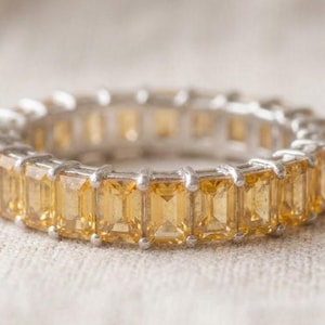 Puede incluir: Un anillo de eternidad con una hilera continua de piedras preciosas de citrino amarillo de corte esmeralda rectangular, engastadas en un marco de metal plateado pulido. Las piedras están dispuestas una al lado de la otra, creando una banda amarilla uniforme.