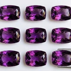 May include: Nine cushion-cut amethyst gemstones in varying shades of purple are displayed. The gemstones are arranged in three rows of three, showcasing their facets and color. A pair of tweezers holds one of the gems, highlighting its clarity and cut.