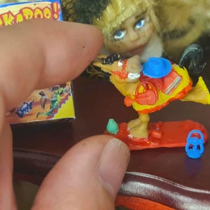 Può includere: Una figurina di un canguro di giocattolo giallo e rosso che cavalca un surf rosso. Il canguro indossa un cappello blu e un giubbotto giallo e rosso. La figurina tiene una borsa rossa. La figurina è su una superficie di legno.