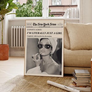 Op de afbeelding: Een ingelijste print die lijkt op een vintage krant, met een zwart-wit afbeelding van een vrouw die lippenstift aanbrengt. De kop luidt "EXPERTS AGREE: I'M LITERALLY JUST A GIRL." De print wordt weergegeven op een tapijt in een lichte kamer.