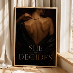 May include: A framed wall art print featuring a dark-toned portrait of a back with a pearl necklace and black lace garment. The text SHE DECIDES is printed in gold capital letters at the bottom, separated by a small gold fleur-de-lis symbol.