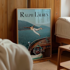 May include: Framed Ralph Lauren advertisement featuring a man diving into the water from a wooden boat. The image has a light blue and brown color scheme, with the brand name in black at the top. The boat's steering wheel and dashboard are visible.