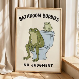 May include: A framed wall art print on a cream background featuring two green frogs. One frog sits on a light blue toilet, while a smaller frog sits on the tank. The text reads BATHROOM BUDDIES at the top and NO JUDGMENT at the bottom.