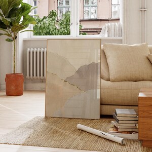 May include: A framed abstract art print featuring a watercolor design in neutral tones of beige, gray, and gold. The artwork is displayed on a beige rug, leaning against a beige sofa. A potted plant and a window are in the background.
