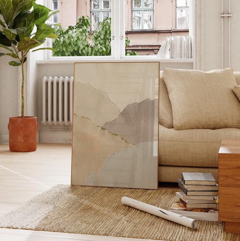 May include: A framed abstract art print featuring a watercolor design in neutral tones of beige, gray, and gold. The artwork is displayed on a beige rug, leaning against a beige sofa. A potted plant and a window are in the background.