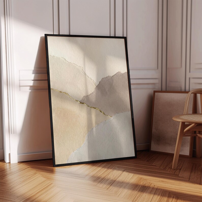 May include: Abstract art print in a black frame. The artwork features a layered design with beige, tan, and gray tones, accented by a gold line. The print is leaning against a white wall and wooden floor, creating a minimalist aesthetic.