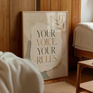 以下が含まれることがあります： 「YOUR VOICE. YOUR RULES.」のテキストがセリフ体で書かれた、額装されたアートプリント。抽象的な形、金箔のアクセント、植物の要素を取り入れたニュートラルな色使いのアートワークです。フレームは明るい木の色です。