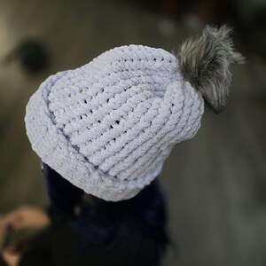 Puede incluir: Un gorro de punto azul claro con un pompón gris esponjoso. El gorro está hecho de un material de punto suave y grueso. El pompón está unido a la parte superior del gorro. El gorro está diseñado para mantener la cabeza caliente.