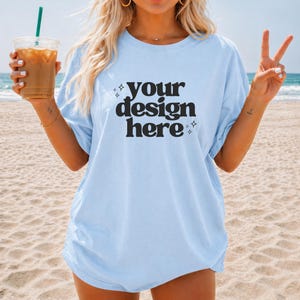 Op de afbeelding: Lichtblauw T-shirt met de tekst "your design here" in een vetgedrukte, zwarte letter. Het shirt heeft een losse pasvorm en lijkt van zacht materiaal te zijn. De foto is genomen op een strand met de oceaan op de achtergrond.