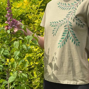 Può includere: Una maglietta verde oliva chiaro con un design botanico dipinto a mano. Il design presenta un gambo marrone con foglie verdi. La maglietta è a maniche corte e realizzata in materiale morbido. Lo sfondo è un cespuglio verde e giallo.