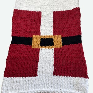 Könnte beinhalten: Eine handgefertigte, grob gestrickte Decke, die dem Anzug des Weihnachtsmanns nachempfunden ist. Die Decke hat einen roten Körper mit schwarzem Gürtel und goldener Schnalle, weißen Besatz und einen weißen oberen Teil. Perfekt für die Weihnachtszeit.