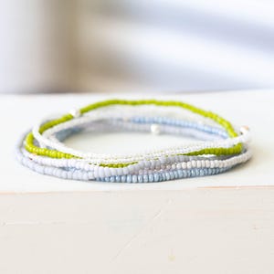 May include: A stack of beaded bracelets in shades of blue, white, and green. The bracelets are made of small, round beads and are arranged in a circular pattern. The bracelets are on a white surface.