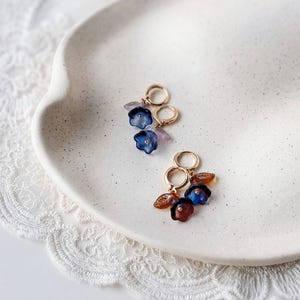 May include: A pair of gold hoop earrings with dangling blue flower charms and small leaf accents. The earrings are displayed on a white, speckled ceramic plate, with a delicate lace trim visible in the background.