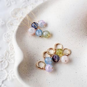 Puede incluir: Pendientes de piedras preciosas en varios colores, incluyendo rosa, azul, verde y morado. Los pendientes se muestran en un plato de cerámica blanco. Los pendientes tienen aros dorados y son adecuados para el uso diario.