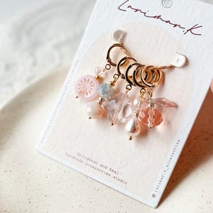 Puede incluir: Un conjunto de marcadores de punto dorados con cuentas y abalorios rosas, transparentes y nacarados. Los marcadores de punto están sujetos a una tarjeta blanca con el texto "Larimar.k" y "Enlighten and Heal. Handmade accessories studio."