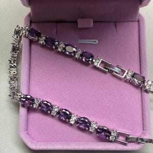 May include: A silver bracelet featuring oval purple gemstones and small white gemstones. The bracelet is presented in a purple velvet jewellery box. The clasp is silver. The bracelet is on a white surface.