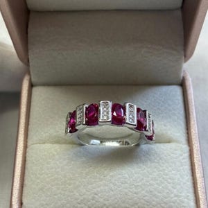 Puede incluir: Un anillo de plata con piedras preciosas rojas ovaladas y pequeños acentos transparentes. El anillo se presenta en una caja de joyería rosa, resaltando su diseño. Las piedras están dispuestas en una fila.