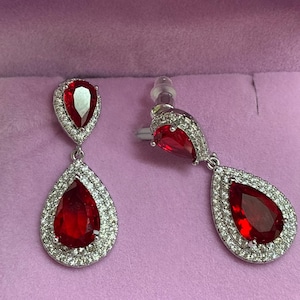 Peut inclure: Une paire de boucles d'oreilles pendantes avec une pierre rouge en forme de poire en haut et une pierre rouge plus grande en forme de poire en bas, entourées d'un halo de petites pierres claires. Monture métallique et fermoirs transparents.