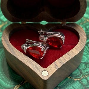 Peut inclure: Boucles d'oreilles en argent avec de grandes pierres rouges ovales. Elles sont présentées dans un écrin en bois en forme de cœur, doublé de velours rouge. La boîte est ouverte.