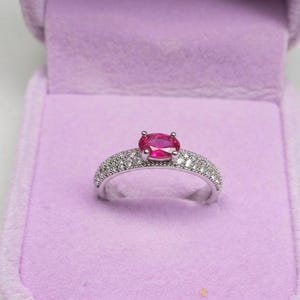 Peut inclure: Une bague en argent avec une grosse pierre précieuse rose vif de coupe ovale au centre. La bande est incrustée de petites pierres précieuses claires. La bague est présentée dans un écrin en velours violet.