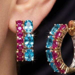Puede incluir: Pendientes dorados con filas alternas de piedras preciosas ovaladas azules y magenta. La vista de cerca resalta el brillo de las gemas y el intrincado engaste.
