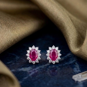 May include: A pair of stud earrings with oval, deep red gemstones encircled by a halo of small, sparkling white stones. The earrings are set against a dark blue surface with a gold fabric in the background.