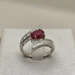 May include: A silver-toned ring featuring a central oval-cut deep red ruby gemstone. The band is split into two bypass-style arms, each channel-set with a row of small, clear baguette-cut diamonds that extend along the sides of the ring shank.