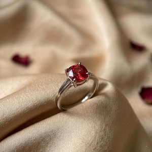 Puede incluir: Un anillo de plata con una gran gema roja de corte cuadrado. El anillo se encuentra sobre un fondo de tela beige con pétalos de rosa dispersos, creando una estética lujosa. La gema es el punto focal.
