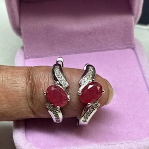 May include: A pair of silver hoop earrings featuring oval-cut red gemstones set in the centre. The curved metal bands are accented with small, clear, sparkling stones along the top and bottom sections of each earring, creating a polished look.