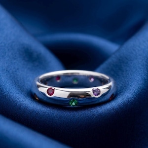 May include: A polished silver band ring featuring multiple small, round, flush-set gemstones in various colours, including red, green, and purple, embedded along the outer surface of the metal band.
