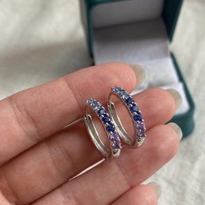 May include: Silver hoop earrings featuring a gradient of blue and purple gemstones. The earrings are presented in an open teal jewellery box. The gemstones transition from light blue to dark blue to purple.