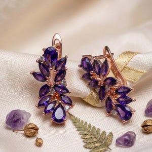 May include: A pair of rose gold earrings featuring a cluster of marquise and pear-cut purple amethyst gemstones arranged in a leaf-like pattern, accented with small clear stones and secured with a hinged lever back closure mechanism.