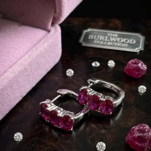 Peut inclure: Boucles d'oreilles créoles en argent ornées de trois pierres roses ovales. Les boucles d'oreilles sont présentées avec des pierres précieuses et de petits diamants sur une surface sombre et réfléchissante. Une boîte à bijoux en velours et une plaque métallique avec "THE BURLWOOD COLLECTION" sont également visibles.