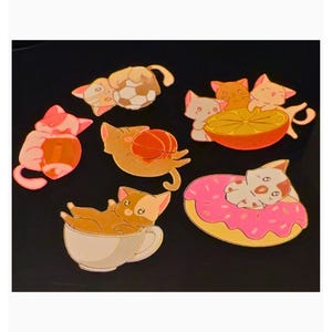 Puede incluir: Una colecci&oacute;n de seis pines de esmalte con gatos en diversas situaciones: uno con un bal&oacute;n de f&uacute;tbol, uno con un bal&oacute;n de f&uacute;tbol americano, uno con un bal&oacute;n de baloncesto, uno en una taza de t&eacute;, uno con un c&iacute;trico y otro sobre un d&oacute;nut rosa.