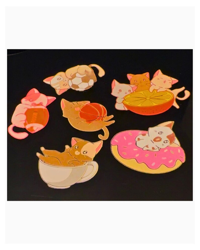Puede incluir: Una colecci&oacute;n de seis pines de esmalte con gatos en diversas situaciones: uno con un bal&oacute;n de f&uacute;tbol, uno con un bal&oacute;n de f&uacute;tbol americano, uno con un bal&oacute;n de baloncesto, uno en una taza de t&eacute;, uno con un c&iacute;trico y otro sobre un d&oacute;nut rosa.