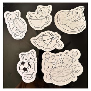 Puede incluir: Una colecci&oacute;n de seis pegatinas troqueladas en blanco y negro con gatos de dibujos animados. Los dise&ntilde;os incluyen un gato con un bal&oacute;n de f&uacute;tbol americano, en una taza, sobre un d&oacute;nut, con un bal&oacute;n de baloncesto, un bal&oacute;n de f&uacute;tbol y una fruta c&iacute;trica.