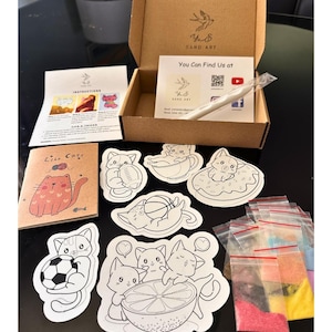 Puede incluir: Un kit de arte con arena que incluye una caja de cart&oacute;n marr&oacute;n, una hoja de instrucciones, un peque&ntilde;o folleto titulado Like Cats y varios contornos adhesivos de gatos. El set contiene un l&aacute;piz blanco y bolsas con arena de colores.