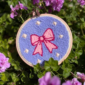 May include: A hand-stitched embroidery hoop featuring a pink bow on a light blue background with white star accents. The embroidery is set in a natural wood hoop, surrounded by green foliage and pink flowers.