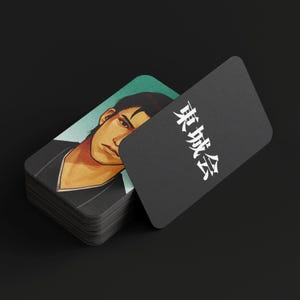Può includere: Una pila di carte nere con angoli arrotondati. Una carta mostra un ritratto di un uomo con capelli scuri e un'espressione seria. Un'altra carta mostra il testo giapponese "東城会" in bianco.