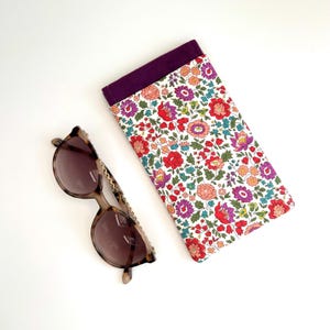 May include: A white and purple floral patterned glasses case with a purple lining. The case is lying on a white surface next to a pair of brown tortoise shell sunglasses with a gold chain.
