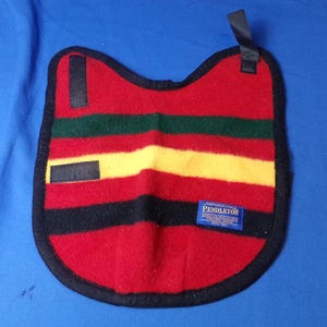 May include: A red, black, green, and yellow striped Pendleton saddle blanket pad. The pad has a black border and a blue label with white text. It has black straps and is on a blue background.