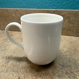 May include: A white ceramic mug with a rounded body and a curved handle. The mug has a smooth, glossy finish and is designed for holding hot or cold beverages. The mug is empty and ready to be filled.