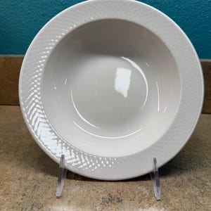 May include: A white ceramic bowl with a textured rim and a smooth, deep center. The rim features a dotted pattern and a subtle chevron design. The bowl is displayed on a clear plastic stand against a teal background.