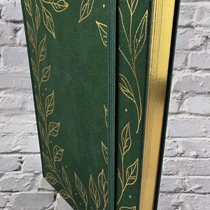 Könnte beinhalten: Ein dunkelgrünes Notizbuch mit festem Einband und weicher Oberfläche, verziert mit goldenen Blattillustrationen auf dem Deckel. Das Notizbuch verfügt über ein passendes grünes Gummiband zum Verschließen und goldfarbene Seitenschnitte.